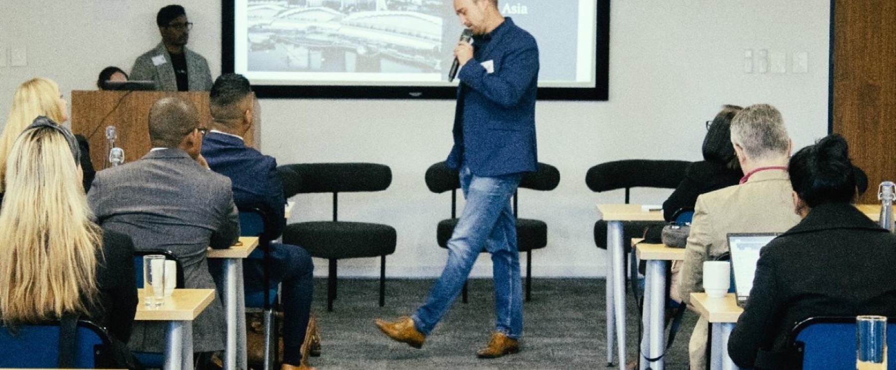 A speaker walking in front of a seated audience at a seminar, beside a projected slide