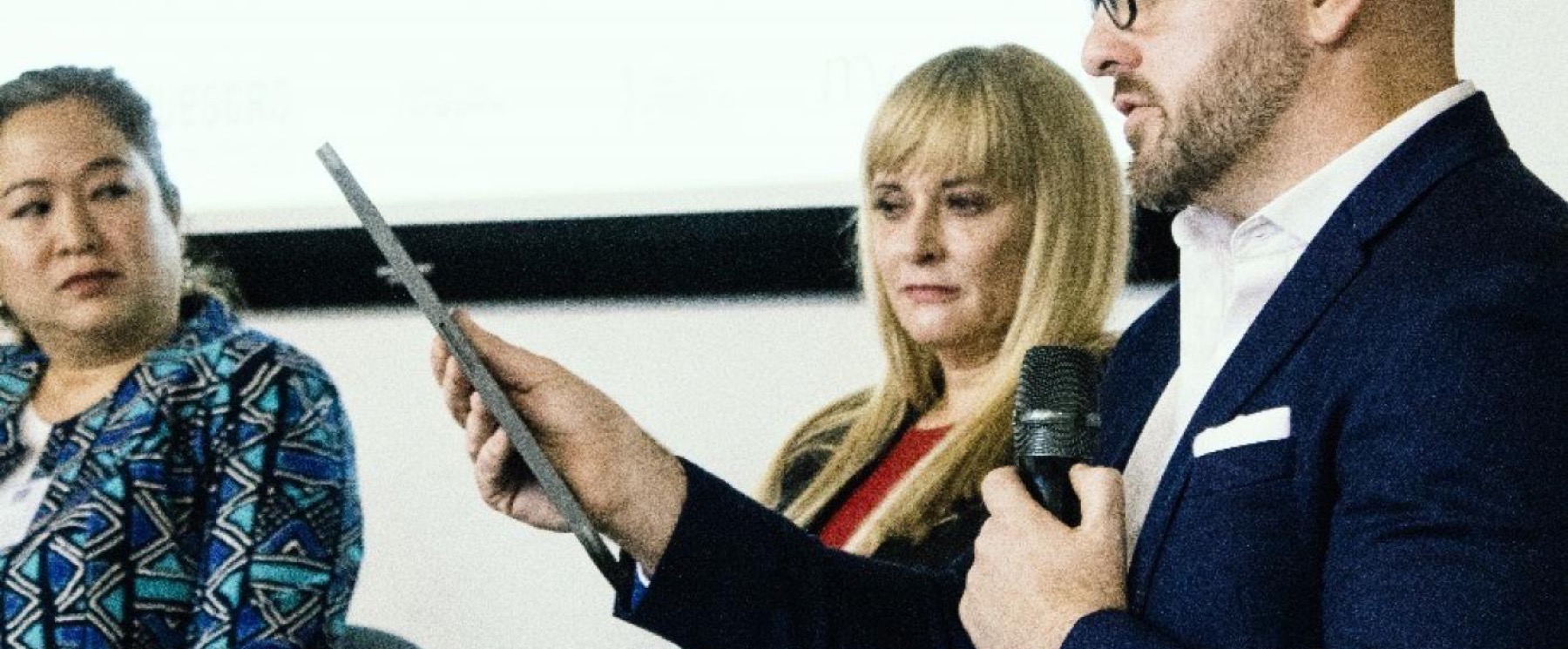 A speaker reading from a card into a hand microphone beside two seated panellists